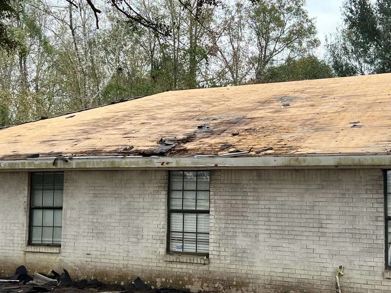 A brick house with its shingles stripped, showing damaged decking along the eave