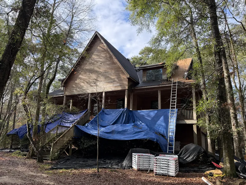 A two-story wood house in the trees with blue tarps over the porch and bundles of new shingles stacked in front