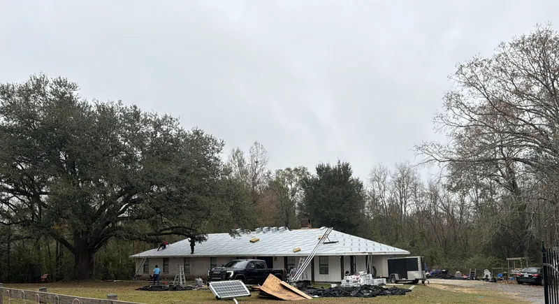 A single-story house under a live oak with its roof dried in, ladders and materials in the yard