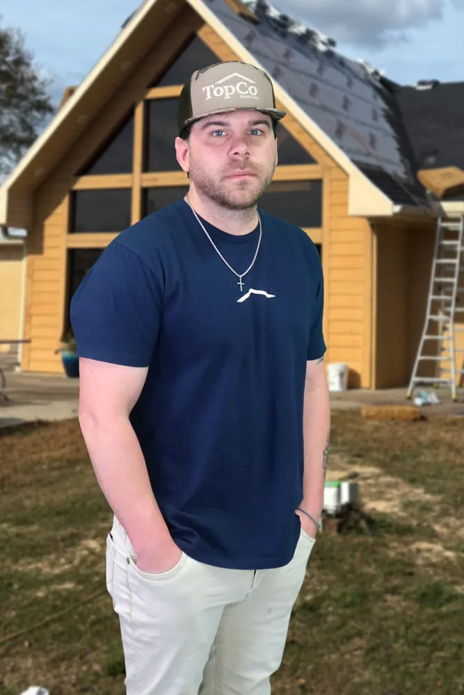 A TopCo team member in a TopCo cap on a job site, with a roof going on behind him